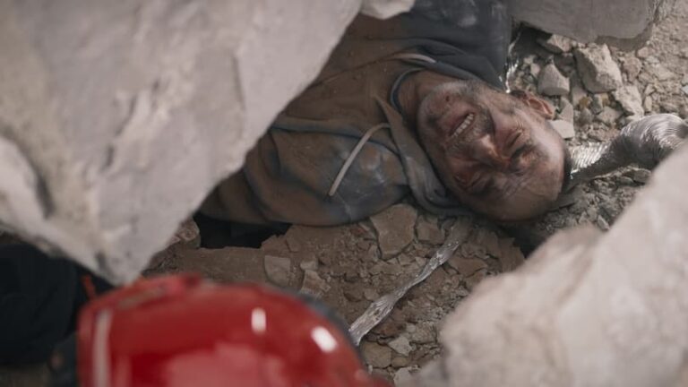 A person is trapped under large debris and appears to be in significant pain, likely due to a catastrophic injury. A red hard hat is visible in the foreground, indicating the presence of rescue efforts. The scene suggests a catastrophic event such as a building collapse.